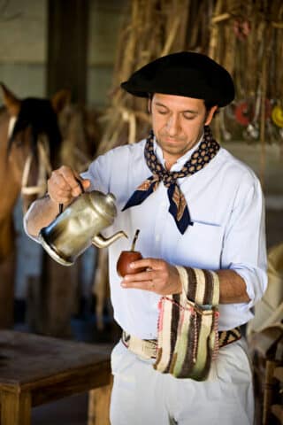 A man in traditional Argentine attire pours a beverage from a metal kettle into a gourd. A horse and rustic equipment can be seen in the background.