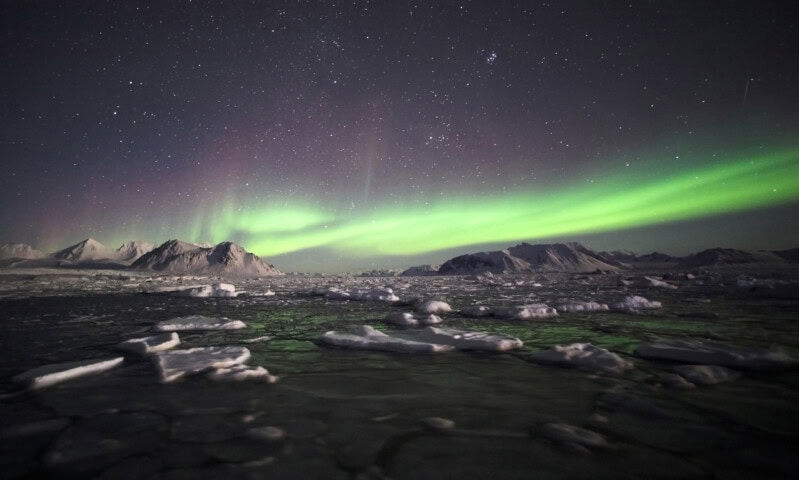 The Northern lights with green and purple hues illuminate the sky over a snowy, mountainous landscape in Greenland, with stars visible and ice floes floating in the foreground, making it a must-visit destination for any travel enthusiast interested in Arctic tourism.