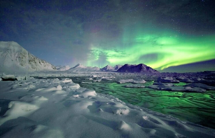 A snowy landscape in Greenland with mountains under a green aurora borealis in the night sky, reflecting on partially frozen water below. This nature scene is a traveler's dream come true.