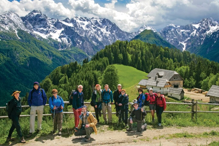A group of hikers poses for a photo on a dirt path with a wooden fence, set against the breathtaking Slovenian mountainous and green forested landscape, complete with several charming alpine buildings in the background.