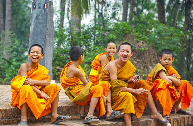 A group of young monks in orange robes sit and laugh on a brick ledge outdoors in Laos.