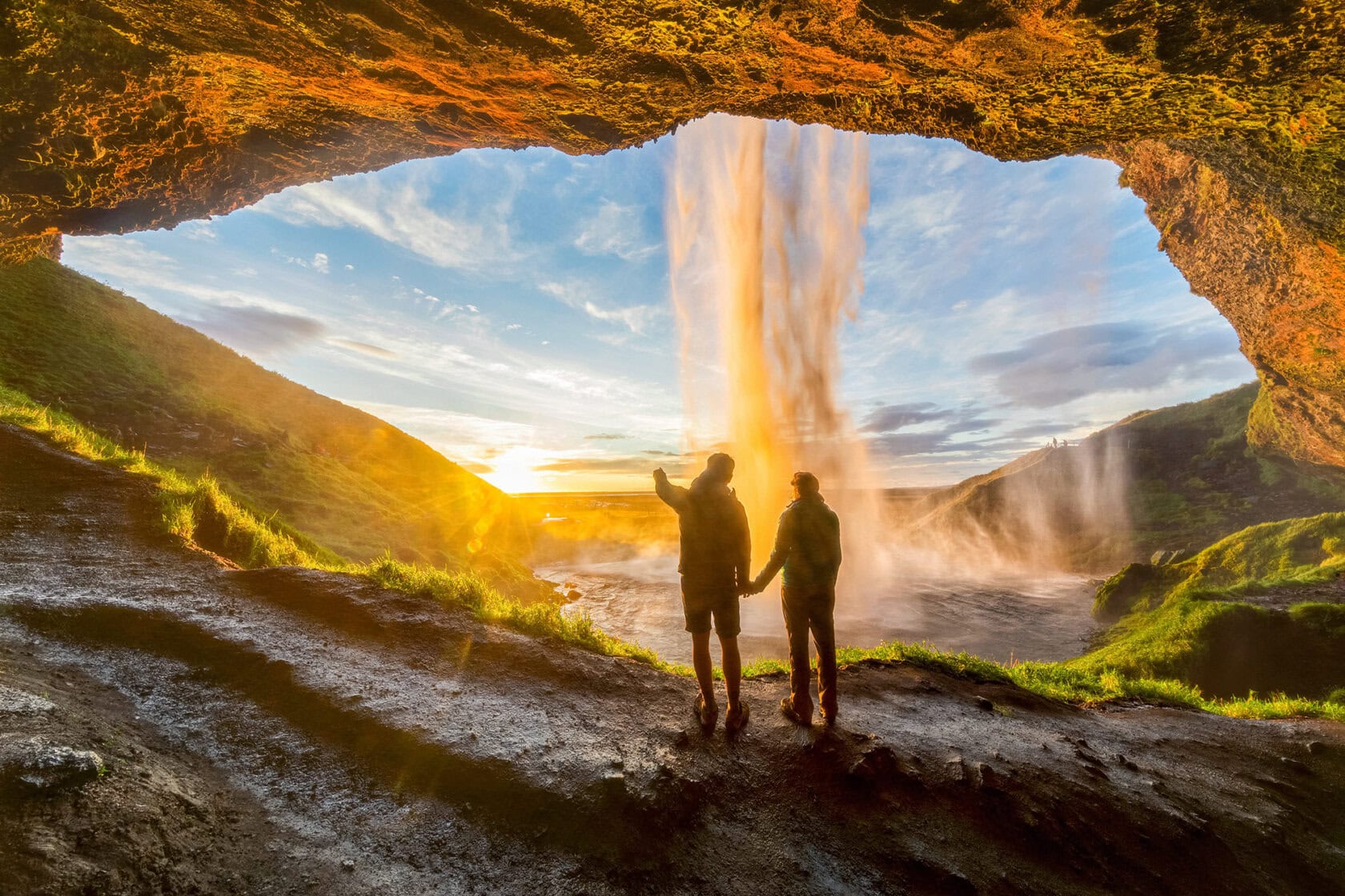 Two people stand in a cave behind a waterfall, holding hands and facing the sunset over a grassy landscape, their trip guided by the promise of adventure.