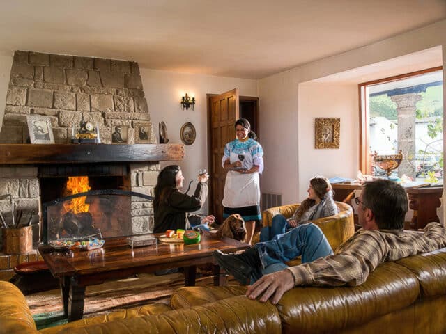 People sitting on sofas around a wooden table in a cozy living room with a stone fireplace, while a woman stands holding a tray, evoking the warmth and charm often found in homes across Ecuador.