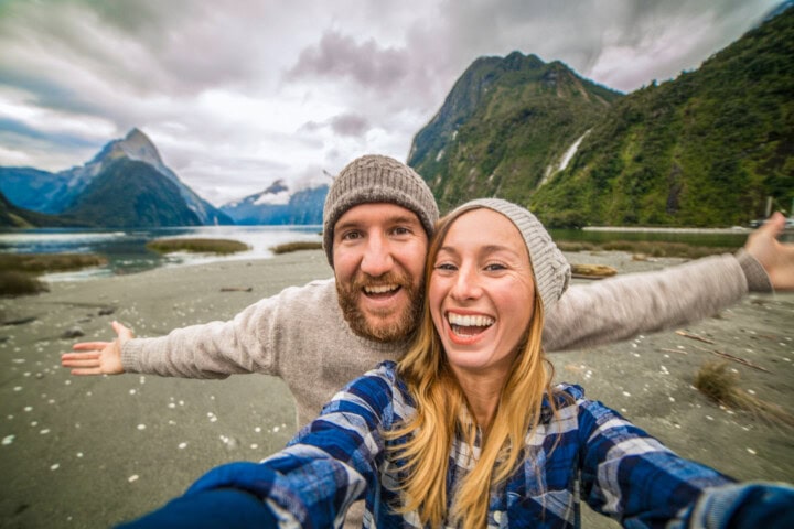 A man and woman in beanies and casual clothes smile while posing with arms outstretched in a scenic New Zealand mountainous area, a river flowing below under a cloudy sky.