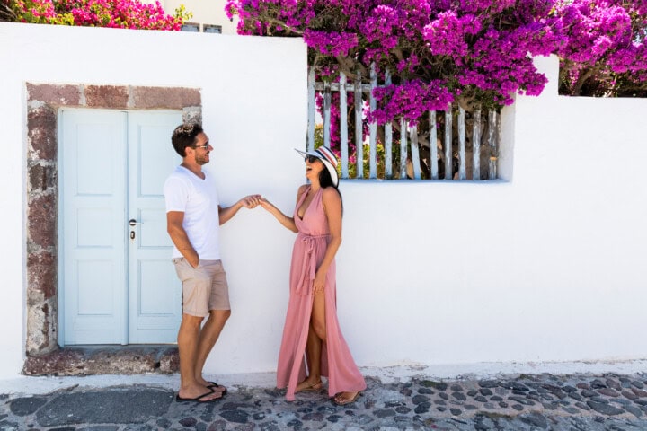 A man in a white shirt and beige shorts and a woman in a pink dress and sun hat stand and smile by a white wall with vibrant purple flowers, capturing the essence of Greece.