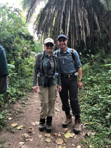 Two travelers wearing hiking gear stand close together on a dirt path in a lush, tropical forest. They are smiling and have their arms around each other, enjoying the adventure.