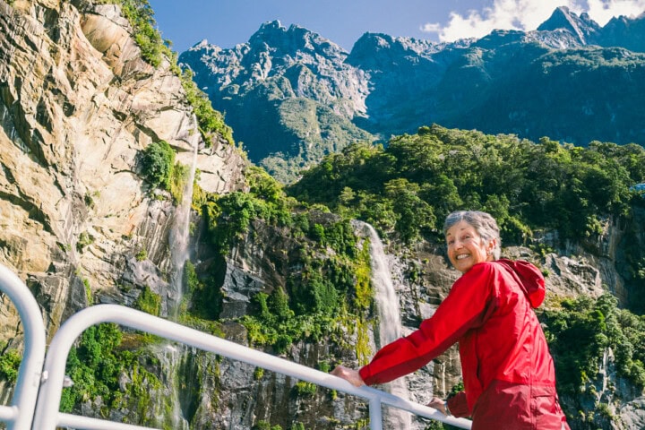 A person wearing a red jacket stands on a railing with waterfalls and a mountainous landscape in the background, capturing the breathtaking beauty of New Zealand.