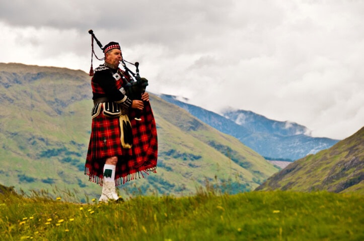 A person dressed in traditional Scottish attire plays bagpipes on a grassy hill with a breathtaking Scotland mountainous landscape in the background, creating an unforgettable travel experience.