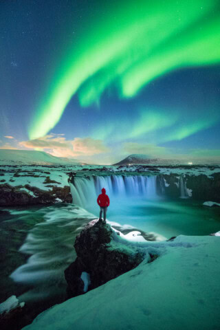 A traveler in a red jacket stands on a snow-covered rock near a waterfall, with the Northern Lights illuminating the night sky above, offering a stunning glimpse into Iceland's natural beauty that beckons tourism.