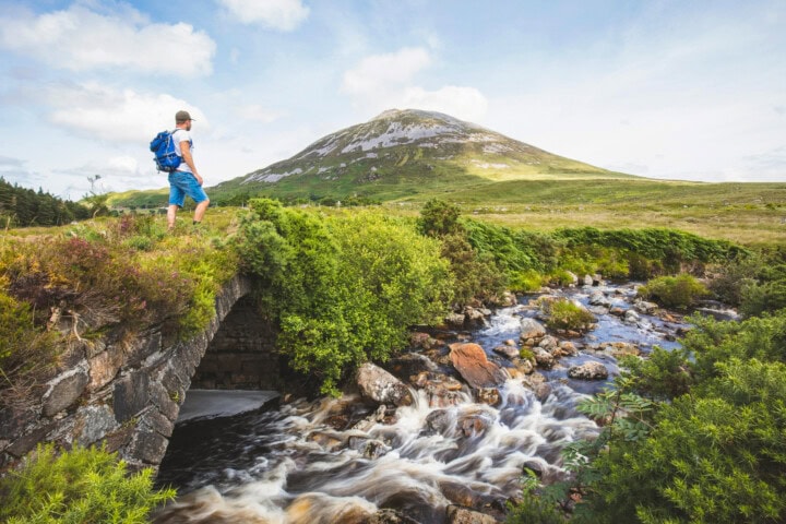 A person with a backpack stands on an old stone bridge above a flowing stream, surrounded by the grassy lands and mountainous landscape of Ireland, all under a partly cloudy sky.