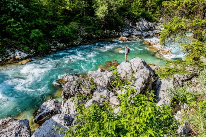 A person stands on large rocks beside a clear, turquoise river surrounded by lush green trees and vegetation.
