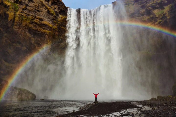 A person stands with arms raised in front of a large waterfall with mist and a vivid rainbow arching across the scene.