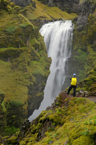 A person with a yellow backpack stands on a moss-covered cliff in Iceland, capturing the majestic waterfall cascading into the rocky gorge, a perfect moment of travel and tourism.