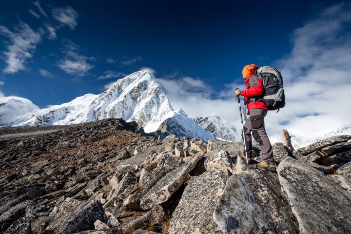 A hiker wearing a red jacket and carrying a large backpack stands on rocky terrain in Nepal, gazing at a snow-covered mountain under a clear blue sky.