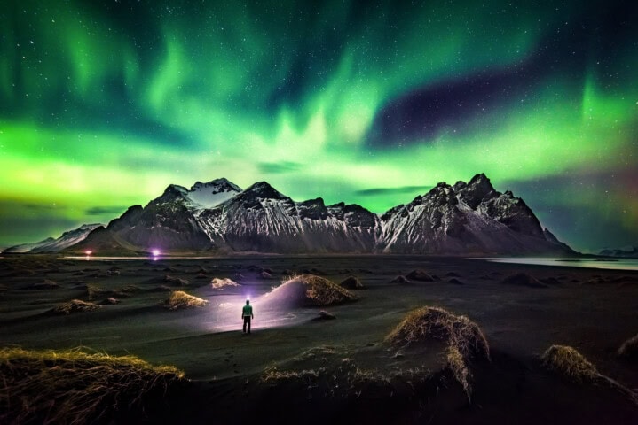 A person stands on a dark landscape in Iceland with mountains in the background, illuminated by the green and purple lights of the northern lights in the night sky—a dream scene that beckons travelers and tourism enthusiasts alike.