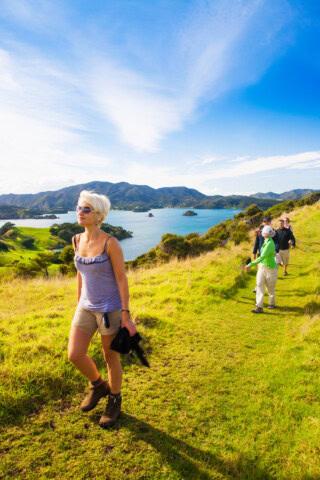 A person in a tank top and shorts walks on a grassy trail near the coast of New Zealand, with three other people in the background. Scenic hills and a blue sky with clouds are in the distance.