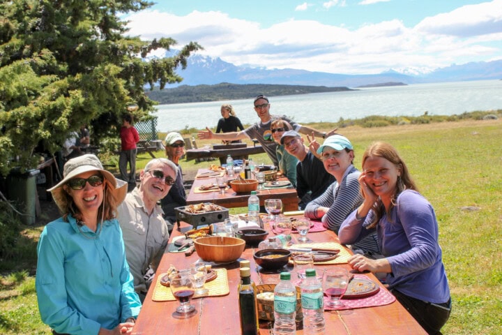 A group of people is sitting at picnic tables outdoors in Argentina, smiling and eating with a scenic lake and mountain backdrop.