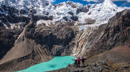 Two people stand on a rocky ledge with arms outstretched, celebrating a 60th birthday while overlooking a turquoise glacial lake surrounded by rugged mountains covered in snow under a blue sky.