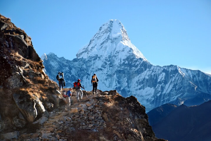 Four hikers trek along a rocky path in Nepal with a snow-capped mountain peak in the background under a clear blue sky.
