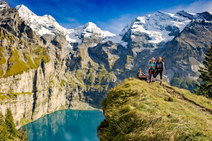 Three hikers are on a grassy cliff overlooking a turquoise alpine lake surrounded by rugged, snow-capped mountains under a clear blue sky, showcasing the breathtaking beauty of Swiss travel.