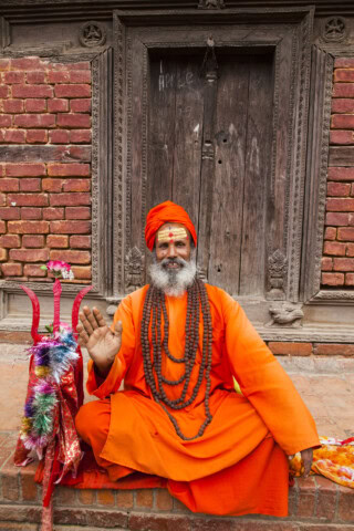 A man dressed in bright orange traditional clothing, adorned with beads and a turban, sits cross-legged and raises one hand in front of an old, intricately carved door in Nepal.