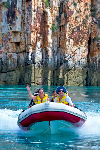 A group of people in yellow life jackets enjoy a boat ride near a rocky cliff in Australia, with one person waving.