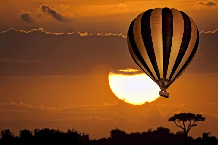 A black-and-white striped hot air balloon floats majestically in front of a large setting sun, with trees and orange skies painting the backdrop—an adventure that promises unforgettable memories.