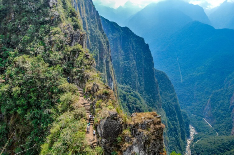 Steep mountain trail with hikers ascending, surrounded by lush green cliffs and a deep valley below, under a partly cloudy sky.