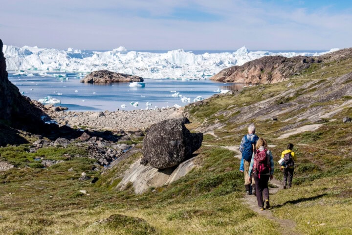 Three hikers on a path in a rocky and grassy landscape in Greenland approach a scenic view of icebergs floating in a bay, with distant snowy peaks under a clear sky.