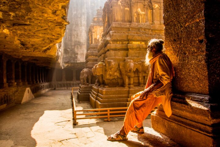 A person dressed in traditional clothing leans against a pillar, looking at ancient rock-cut architecture with carvings of elephants. Sunlight filters through the space, creating a warm ambiance, encapsulating the spirit of an unforgettable adventure.
