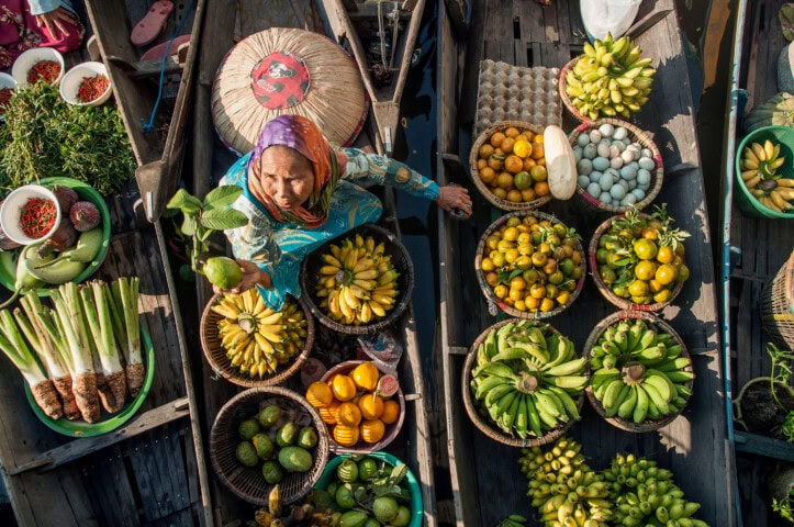 A woman in a boat at a Malaysian floating market sells various fruits and vegetables, including bananas, limes, and eggs. Fresh produce is displayed in baskets around her.