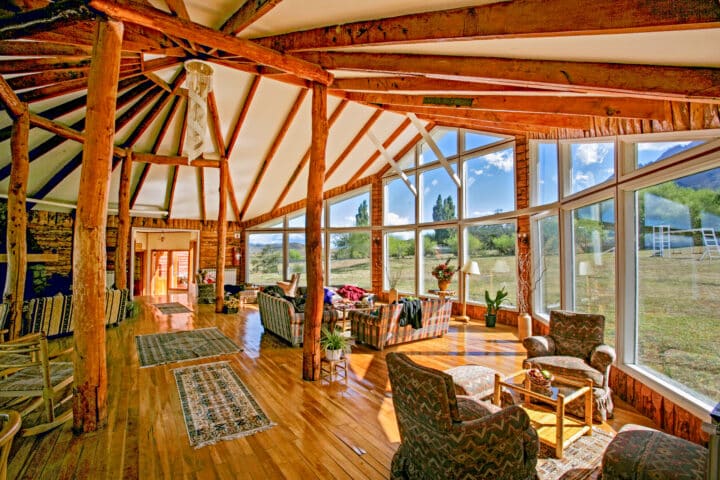 A spacious, sunlit room with large windows and exposed wooden beams. It features several seating areas with armchairs and couches, a wood floor, and views of the Chilean outdoors.