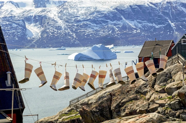 A row of traditional clothing, including socks, hangs on a line to dry with an icy ocean and the snow-capped mountains of Greenland in the background – a picturesque moment that captures the essence of travel and tourism.