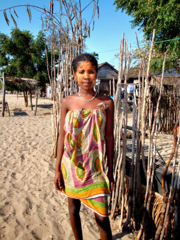 A woman stands in a sandy area, wearing a colorful patterned dress with a white necklace, reminiscent of styles from The Eighth Continent. Sparse wooden structures and trees are visible in the background.
