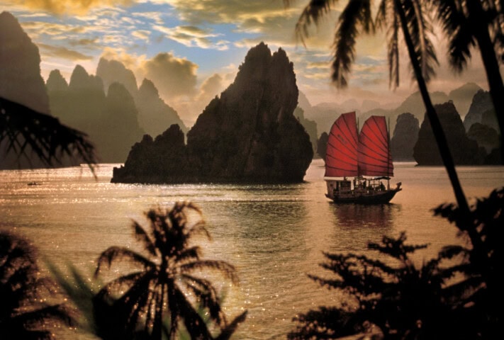 A traditional boat with red sails navigates a calm water body in Vietnam, surrounded by rocky outcrops and silhouetted palm trees under a partly cloudy sky at sunset.