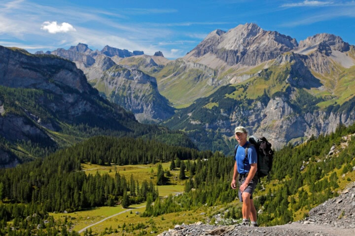 A person wearing a blue shirt and shorts stands on a mountainous trail in Switzerland, with a scenic view of high peaks, valleys, and forests in the background under a clear blue sky.