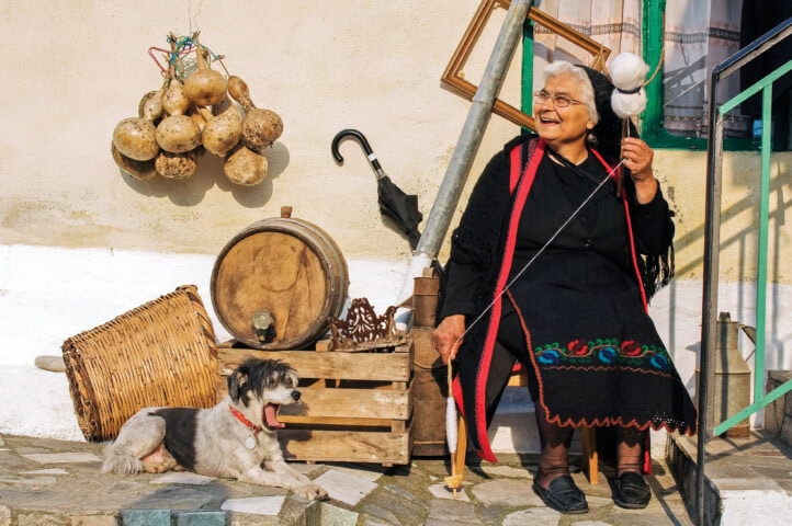 An elderly woman spins wool while sitting outside on a sunny day in Greece, with gourds hanging on the wall and a small dog resting nearby. Various household items, including a barrel and baskets, are around her.