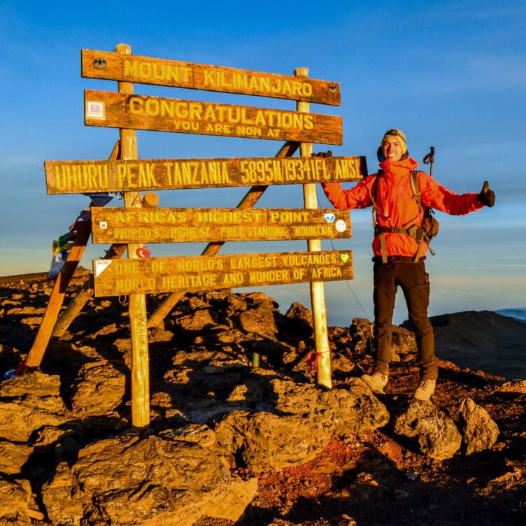 A person in hiking gear stands next to the Mount Kilimanjaro summit sign at Uhuru Peak, capturing moments not to miss in 2026, with rocky ground and a bright sky in the background.