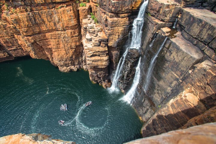 Aerial view of a tall waterfall cascading into a pool surrounded by steep rocky cliffs in Australia, with a few boats creating circular patterns on the water.