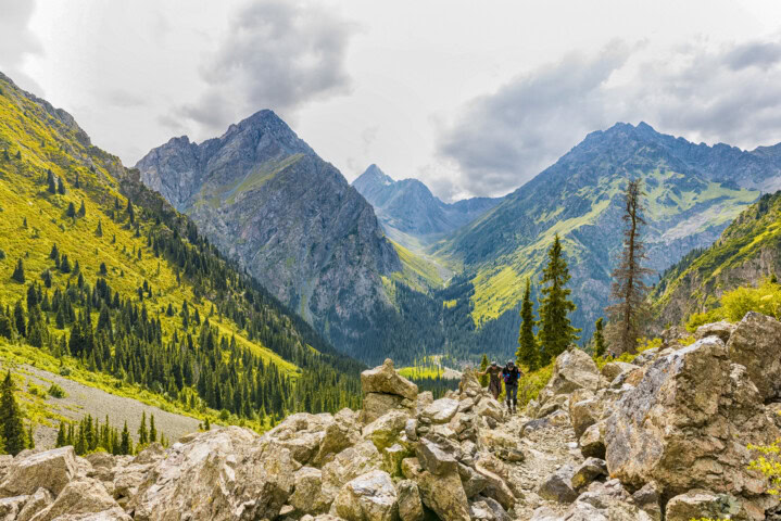 A hiker walks along a rocky path in a mountain valley in Kyrgyzstan, with steep, forested slopes and cloudy skies.