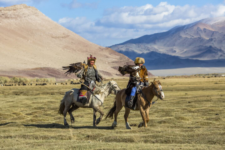 Two individuals riding horses carry eagles on their arms in an open, grassy landscape with the stunning mountains of Kyrgyzstan in the background.