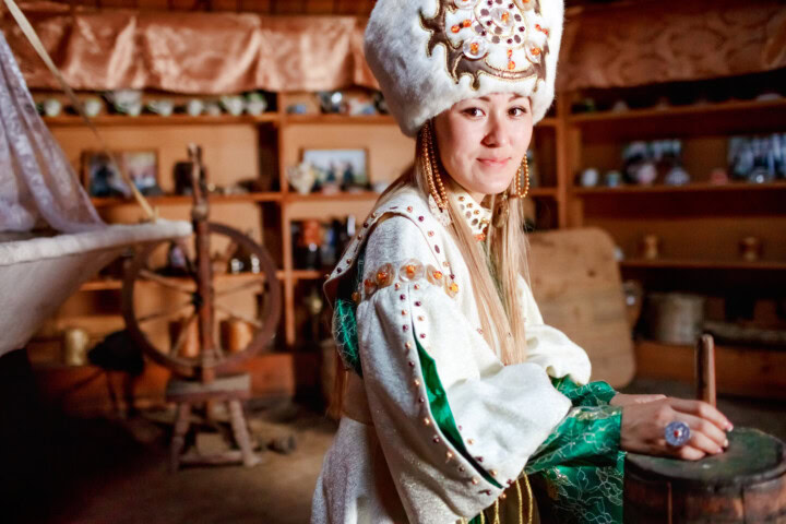 A person in traditional attire with a white fur hat stands in a rustic room reminiscent of Kyrgyzstan, with a spinning wheel and wooden shelves in the background.