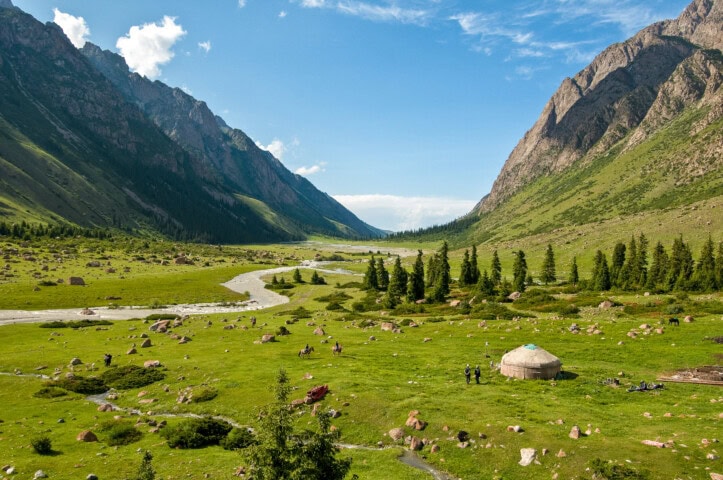 A wide valley in Kyrgyzstan with lush green grass, a river winding through it, and a yurt near the center. The scene is surrounded by tall, rocky mountains under a blue sky with a few clouds.