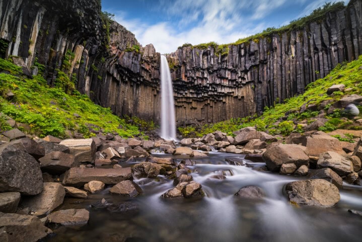 A waterfall cascades from a basalt cliff into a rocky pool surrounded by greenery under a cloudy sky, creating an idyllic scene that epitomizes the natural beauty of Iceland, making it a must-visit spot for tourism and travel enthusiasts.