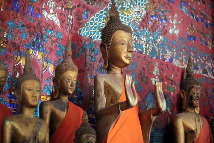 Several Buddha statues in various postures, draped in orange robes, stand in front of a colorful, intricate mural in Laos.