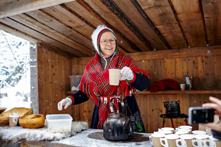 A person wearing traditional attire serves a hot beverage from a kettle at an outdoor wooden stall, reminiscent of Finland, with cups laid out on the counter.