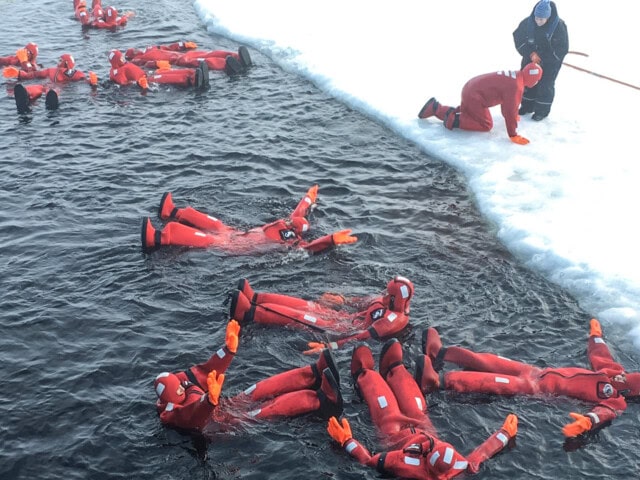 Individuals in red survival suits float in icy water next to an ice shelf in Finland, while a person in warm clothing observes and assists from the edge.