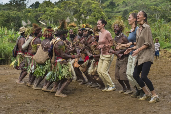 A group of indigenous people in traditional attire dance with four women in casual clothes outdoors, surrounded by the lush greenery of Papua New Guinea.