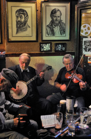 Three elderly men in a dimly lit room, playing traditional musical instruments beneath framed portraits and various decorations on the walls, evoke the soulful rhythms of Ireland.