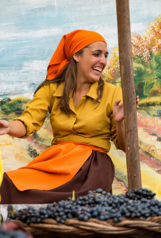 A woman dressed in a yellow and brown outfit with an orange headscarf smiles while sitting next to a basket of grapes in front of a colorful mural, evoking the vibrant spirit of Portugal.